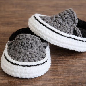 Op de afbeelding: Een paar handgehaakte babyschoentjes in sneakerstijl. De schoenen hebben grijze bovenkanten, witte zolen met een zwarte streep en zwarte binnenvoering. De schoenen worden getoond op een houten oppervlak.