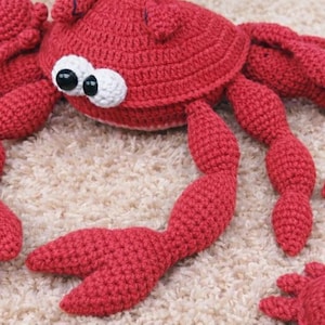 Puede incluir: Un cangrejo rojo hecho a mano con ganchillo, con grandes ojos saltones y múltiples patas. El cangrejo está sobre una alfombra beige. El cuerpo del cangrejo tiene forma redondeada y sus patas están detalladas con un patrón texturizado.