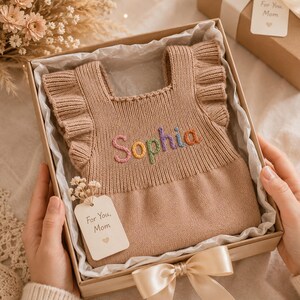Op de afbeelding: Een beige gebreid mouwloos topje met ruches op de schouders, voorzien van de naam Sophia geborduurd in meerkleurig garen op de borst. Het kledingstuk zit in een kartonnen geschenkdoos bekleed met wit vloeipapier en bevat een klein label.
