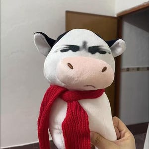 Könnte beinhalten: Ein Plüschkuhspielzeug mit weißem Körper, schwarzen Ohren und Markierungen sowie einer rosa Nase. Die Kuh trägt einen roten Strickschal. Das Spielzeug wird von einer Hand gehalten.