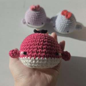 Könnte beinhalten: Handgefertigte Wal-Plüschtiere aus Häkelarbeit in Rosa, Lavendel und Hellblau. Der rosa Wal ist im Fokus, gehalten in einer Hand, mit weißem Bauch und schwarzen Augen. Die anderen Wale haben kleine Schleifen. Weiche Spielzeuge für Kinder.