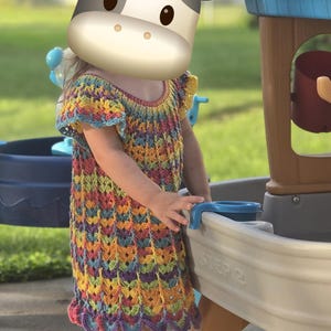 May include: A short-sleeved dress featuring a multi-colored crochet pattern with repeating shell-like stitches in shades of yellow, orange, blue, and purple. The garment has a round neckline and a scalloped hemline, showcasing a textured, open-work design.