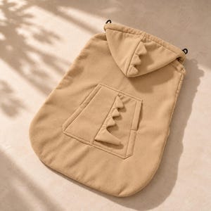 May include: A tan fleece pet hoodie featuring a hood with three small triangular spikes and a central pocket on the back adorned with a vertical row of four matching triangular spikes. The garment includes two black toggles at the neckline.