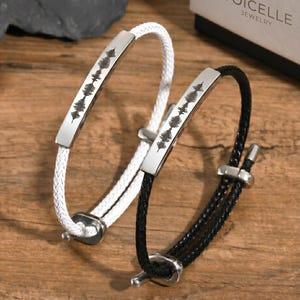 May include: Two bracelets, one white and one black, with a silver metal accent featuring a sound wave design. The bracelets are displayed on a wooden surface. The brand name "Voicelle Jewelry" is visible on a box in the background.