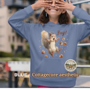 May include: Blue hoodie with a squirrel graphic and the word "Yay!". The design includes acorns and the text "Cottagecore aesthetic" and "BLUE". The hoodie has a front pocket and a "Cottagecore" logo.