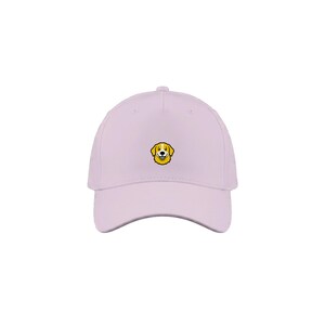 Puede incluir: Gorra de béisbol rosa claro con visera curva. La gorra presenta un pequeño perro de dibujos animados bordado con detalles amarillos, negros y blancos en la parte delantera. El sombrero es de color liso y tiene un diseño clásico de seis paneles.
