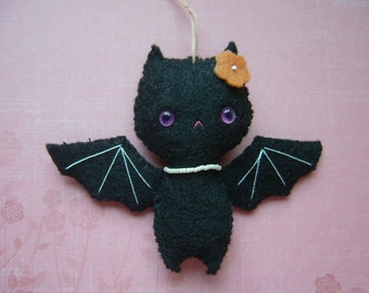 May include: A black felt bat with white stitching for wings and a small orange felt flower on its head. The bat has purple eyes and a white beaded necklace.