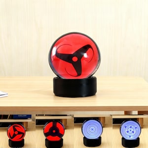 May include: A red and black decorative light with a circular design, set on a black base. The light is spherical and features a three-pronged symbol. The base is cylindrical. Additional lights with different designs are visible.