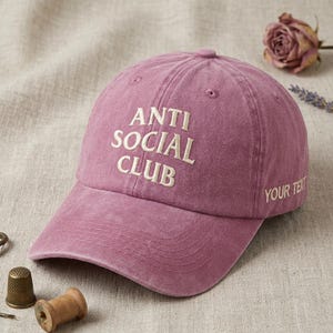 Peut inclure: Casquette de baseball rose poussi&eacute;reux avec les mots "ANTI SOCIAL CLUB" brod&eacute;s en blanc. La phrase "YOUR TEXT" est brod&eacute;e sur le c&ocirc;t&eacute;. La casquette est en tissu doux et textur&eacute;.