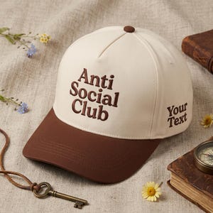 Peut inclure: Casquette de baseball cr&egrave;me et marron avec l'inscription brod&eacute;e "Anti Social Club" sur le devant et "Your Text" sur le c&ocirc;t&eacute;. La casquette a une visi&egrave;re marron et un bouton marron sur le dessus.