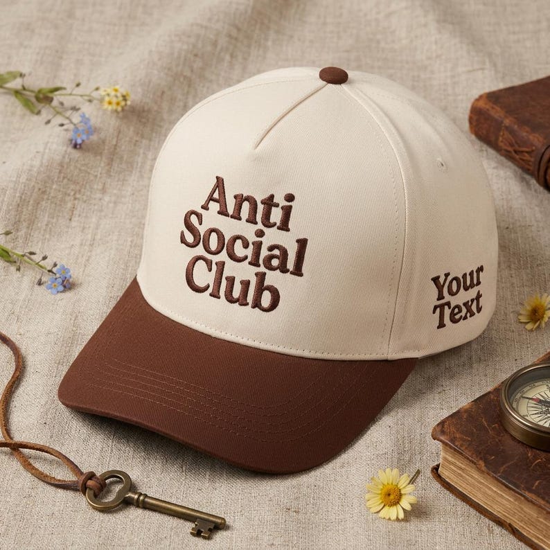 Peut inclure: Casquette de baseball cr&egrave;me et marron avec l'inscription brod&eacute;e "Anti Social Club" sur le devant et "Your Text" sur le c&ocirc;t&eacute;. La casquette a une visi&egrave;re marron et un bouton marron sur le dessus.