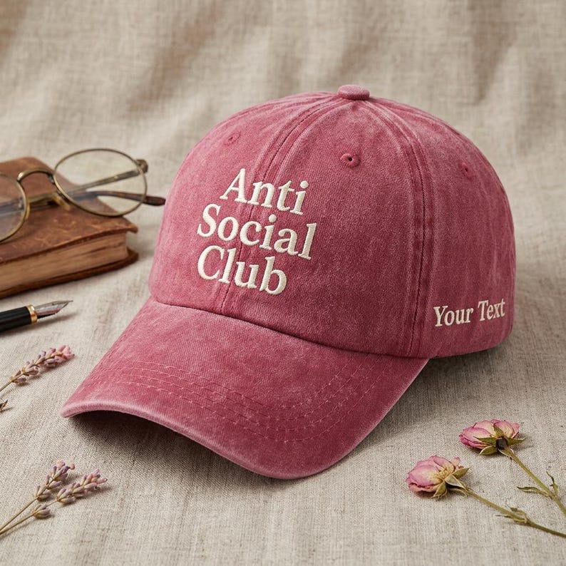 Peut inclure: Casquette de baseball rose fan&eacute; avec les mots "Anti Social Club" brod&eacute;s en blanc sur le devant. L'expression "Your Text" est brod&eacute;e sur le c&ocirc;t&eacute;. La casquette est pos&eacute;e sur une surface de ton neutre.