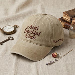 Peut inclure: Casquette de baseball beige avec le texte brod&eacute; "Anti Social Club" en marron. La casquette porte &eacute;galement le texte "Your Text" brod&eacute; sur le c&ocirc;t&eacute;. Une montre de poche vintage, une cl&eacute; et une petite bouteille sont en arri&egrave;re-plan.