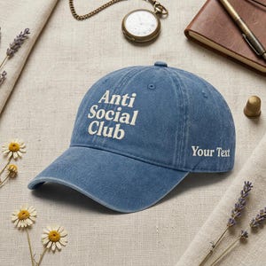 Peut inclure: Casquette de baseball bleu avec le texte blanc "Anti Social Club" et "Your Text". La casquette a une visi&egrave;re incurv&eacute;e et est faite d'un tissu doux. En arri&egrave;re-plan, une montre de poche, un carnet et des fleurs.