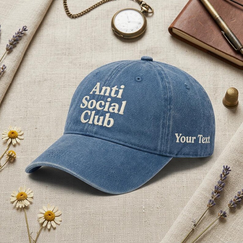 Peut inclure: Casquette de baseball bleu avec le texte blanc "Anti Social Club" et "Your Text". La casquette a une visi&egrave;re incurv&eacute;e et est faite d'un tissu doux. En arri&egrave;re-plan, une montre de poche, un carnet et des fleurs.