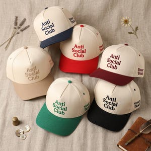 Peut inclure: Casquettes de baseball cr&egrave;me avec des visi&egrave;res color&eacute;es et le texte "Anti Social Club" en diff&eacute;rentes couleurs. Chaque chapeau porte les mots "Your Text" sur le c&ocirc;t&eacute;. Les visi&egrave;res sont bleu marine, rouge, bordeaux, vert, noir et beige.