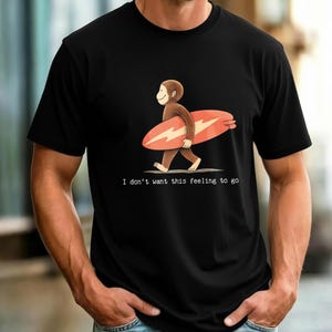 May include: A black short-sleeved t-shirt featuring a graphic of a brown monkey carrying a coral-colored surfboard with a white lightning bolt design. Below the illustration, the text reads, I don't want this feeling to go.