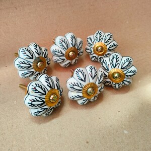 May include: A set of five flower-shaped ceramic knobs featuring a white base with black leaf-like patterns and a central gold-toned metal hardware piece. Each knob has a scalloped edge and a rounded, petal-inspired design with a metallic center.