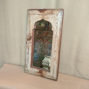 May include: A rectangular wall mirror featuring a weathered, light-coloured wooden frame with a distinct arched top design. The mirror glass reflects a portion of a dark, ornate wooden cabinet with green and blue decorative circular patterns.