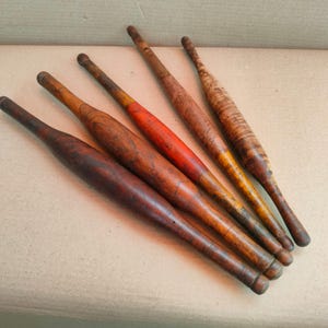 May include: A collection of five vintage wooden rolling pins arranged diagonally. Each pin features a distinct finish, including dark brown, natural wood grain, and one with a vibrant red painted center section, all showing signs of wear.