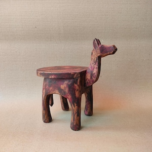 Pode incluir: Um pequeno banco de madeira artesanal com um design único em forma de animal. O banco apresenta um assento redondo e quatro pernas, com uma cabeça e pescoço esculpidos. Pintado em tons de vermelho e castanho, tem um aspeto rústico. Altura: cerca de 25 cm.