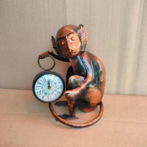 May include: A bronze-colored metal monkey figurine holding a small, round clock. The clock face is white with black numerals and hands. The monkey has detailed features and is posed in a seated position, with its tail curled.
