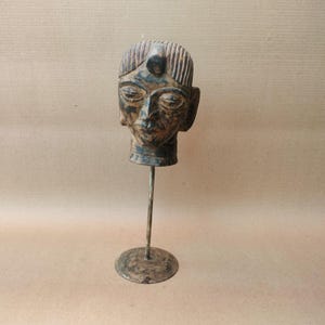 May include: A weathered, wooden sculpture of a human head mounted on a metal stand. The head features carved details, including closed eyes, a prominent nose, and a stylized hairstyle. The sculpture has a dark, aged appearance.