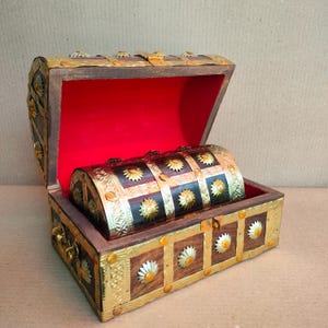 May include: A wooden chest with a hinged lid, featuring brass-colored metal trim and decorative sunburst studs. The interior is lined with bright red fabric. A smaller, matching wooden chest with similar brass accents sits inside the open main chest.