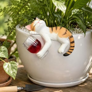 Puede incluir: Una figura de gato naranja y blanco durmiendo sobre una maceta de cerámica blanca. El gato sostiene una copa de vino llena de líquido rojo. La maceta contiene plantas verdes y está sobre una superficie de madera.