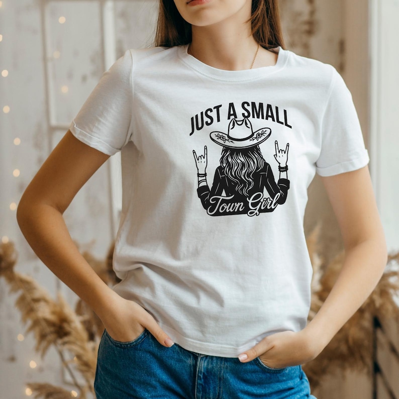 K&ouml;nnte beinhalten: Wei&szlig;es T-Shirt mit schwarzer Grafik, die eine Frau mit Cowboyhut und dem Text "JUST A SMALL Town Girl" zeigt. Das Design beinhaltet Rock'n'Roll-Handgesten. Das Shirt ist aus weichem Material.