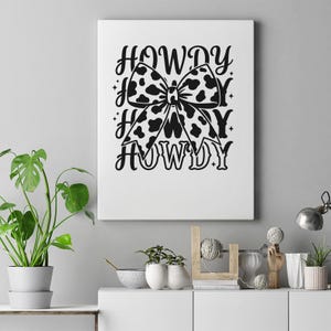 K&ouml;nnte beinhalten: Ein wei&szlig;er Leinwanddruck mit einer schwarz-wei&szlig;en Kuhfell-Schleife und dem Wort "HOWDY" in einer dekorativen Schriftart. Der Bogen ist das zentrale Designelement, mit "HOWDY" dar&uuml;ber und darunter wiederholt.