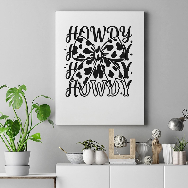 K&ouml;nnte beinhalten: Ein wei&szlig;er Leinwanddruck mit einer schwarz-wei&szlig;en Kuhfell-Schleife und dem Wort "HOWDY" in einer dekorativen Schriftart. Der Bogen ist das zentrale Designelement, mit "HOWDY" dar&uuml;ber und darunter wiederholt.