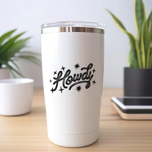 K&ouml;nnte beinhalten: Ein wei&szlig;er Isolierbecher mit transparentem Deckel. Der Becher hat das Wort "Howdy" in schwarzer Schrift, umgeben von schwarzen Sternen. Der zylindrische Becher steht auf einer Holzoberfl&auml;che.