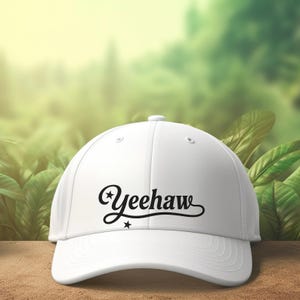 K&ouml;nnte beinhalten: Eine wei&szlig;e Baseballkappe mit dem Wort "Yeehaw" in schwarzer Schrift. Die Kappe hat einen gebogenen Schirm und einen Knopf oben. Der Hintergrund ist ein verschwommenes Bild von gr&uuml;nem Laub.