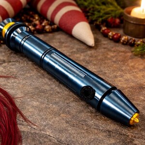 May include: A blue metallic lightsaber prop with gold and black accents. The prop is cylindrical with a button and a silver stripe. The background includes holiday decorations, a lit candle, and a red and white striped item.
