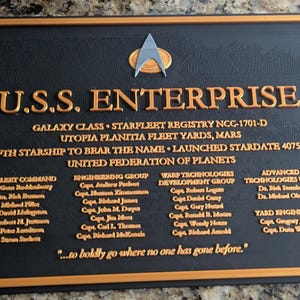 May include: A rectangular black plaque with a gold border and gold lettering. It features the text U.S.S. ENTERPRISE, Starfleet details, crew names, and the quote ...to boldly go where no one has gone before. A silver Starfleet insignia is at the top.