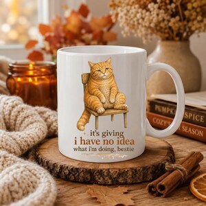 May include: White ceramic mug featuring an orange tabby cat sitting on a wooden chair. The mug has the text "it's giving i have no idea what I'm doing, bestie". The mug is on a wooden coaster with fall-themed decor.