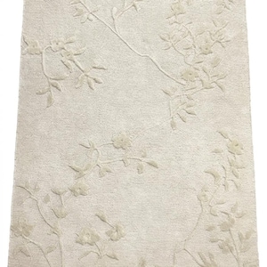 Puede incluir: Alfombra rectangular color crema con un diseño floral en relieve. La alfombra presenta un patrón repetido de flores y hojas estilizadas, creando un aspecto texturizado y elegante. El diseño está en relieve, agregando profundidad al fondo suave y neutro.