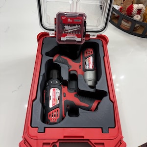 Könnte beinhalten: Ein roter und schwarzer Milwaukee Packout Werkzeugkoffer mit einem transparenten Deckel, der geöffnet ist, um einen Bohrer und einen Schlagschrauber zu zeigen. Eine kleinere rote Hülle mit Milwaukee-Zubehör befindet sich im Inneren.