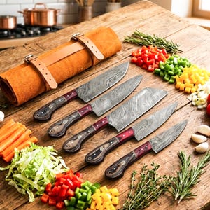 Könnte beinhalten: Ein Set aus fünf Küchenmessern mit Damaststahlklingen und dunkelbraunen Holzgriffen mit roten Akzenten, angeordnet neben einer beigen Leder-Rolltasche. Die Messer liegen auf einer Holzoberfläche neben gehacktem Gemüse und frischen Kräutern.