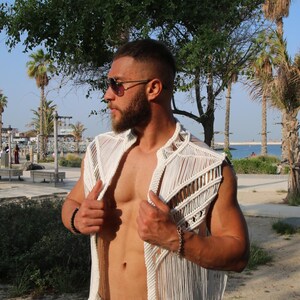 Pode incluir: Um colete branco sem mangas em macramé com um design geométrico. O colete é aberto na frente e tem franjas na parte inferior. A pessoa está usando óculos de sol e uma pulseira. O fundo inclui palmeiras e um céu ensolarado.