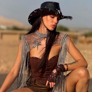 May include: A black cowboy hat with silver rings and a skull emblem, paired with a silver metallic fringe shoulder piece. The outfit includes a brown lace-up corset top, brown shorts, and brown fingerless leather gloves with studs.