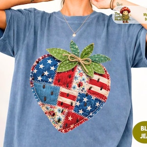 Pode incluir: Camiseta azul jeans com um design patriótico de morango. O morango é feito de retalhos com estrelas, listras e estampas florais. A camiseta tem mangas curtas e decote redondo. O texto "The Lucky Mushroom" e "100% My Original Design" são visíveis.