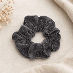 Könnte beinhalten: Ein dunkelgraues Samt-Haargummi, ein Haaraccessoire, liegt auf einem neutralfarbenen Stoff. Das Haargummi ist rund und gerafft, mit einem weichen, strukturierten Aussehen. Zarte, getrocknete Blumen sind in der oberen linken Ecke zu sehen.