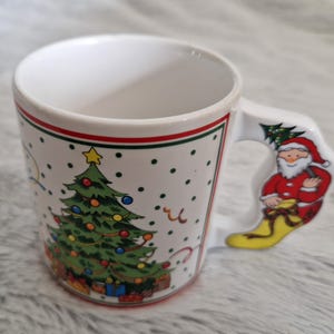 May include: White ceramic Christmas mug with a green Christmas tree and red and white Santa Claus on the handle. The mug has a red and green trim around the top.