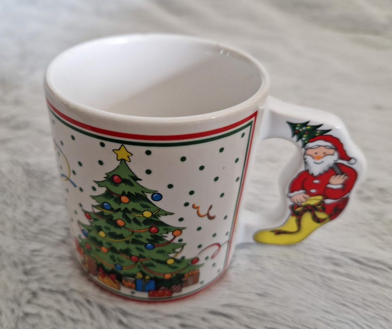 May include: White ceramic Christmas mug with a green Christmas tree and red and white Santa Claus on the handle. The mug has a red and green trim around the top.