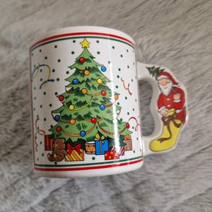 May include: White ceramic Christmas mug with a green Christmas tree, red and white presents, a brown teddy bear, and a Santa Claus holding a gift on the handle.