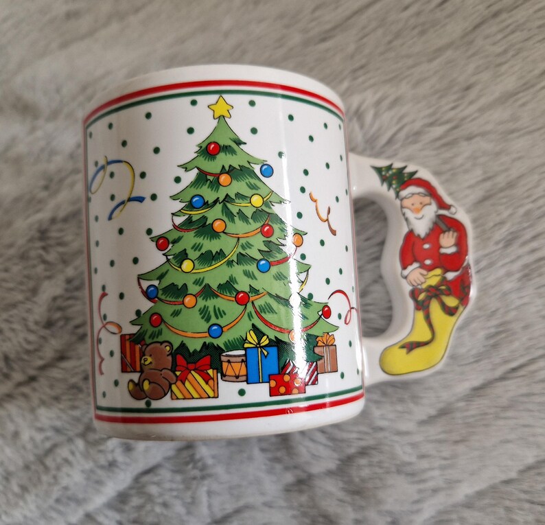 May include: White ceramic Christmas mug with a green Christmas tree, red and white presents, a brown teddy bear, and a Santa Claus holding a gift on the handle.