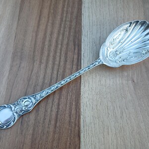 May include: A silver-toned serving spoon featuring an ornate, embossed handle with intricate scrollwork and a shell-shaped bowl. The bowl displays a detailed floral motif at the base of the fluted design, extending into a decorative stem.
