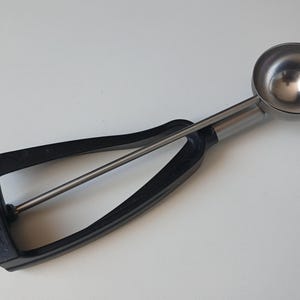 May include: A stainless steel ice cream scoop featuring a black plastic handle with a squeeze-release mechanism. The tool has a polished metal bowl and a black frame that connects to the handle, designed for portioning frozen desserts.